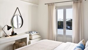 A bedroom with a bed, a bedside table with a lamp, a mirror, and a window with curtains. - Skylira Suite, Partial Sea View, Private Pool or Jacuzzi, Furnished Terrace, Oia (Santorini)