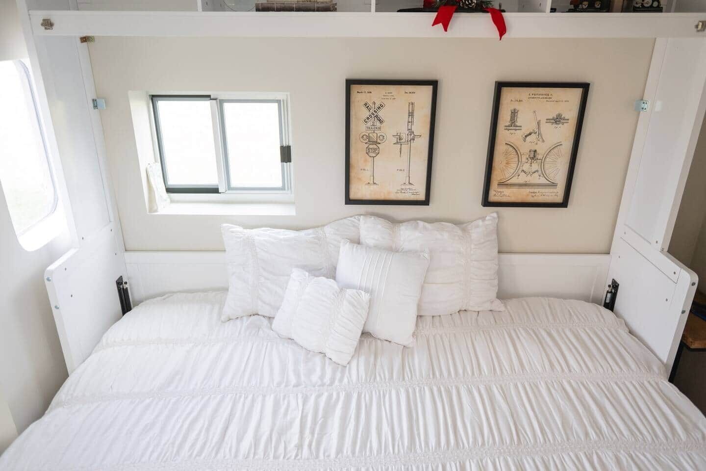 A bed with white bedding and pillows, a small window, and two framed vintage technical drawings on the wall.