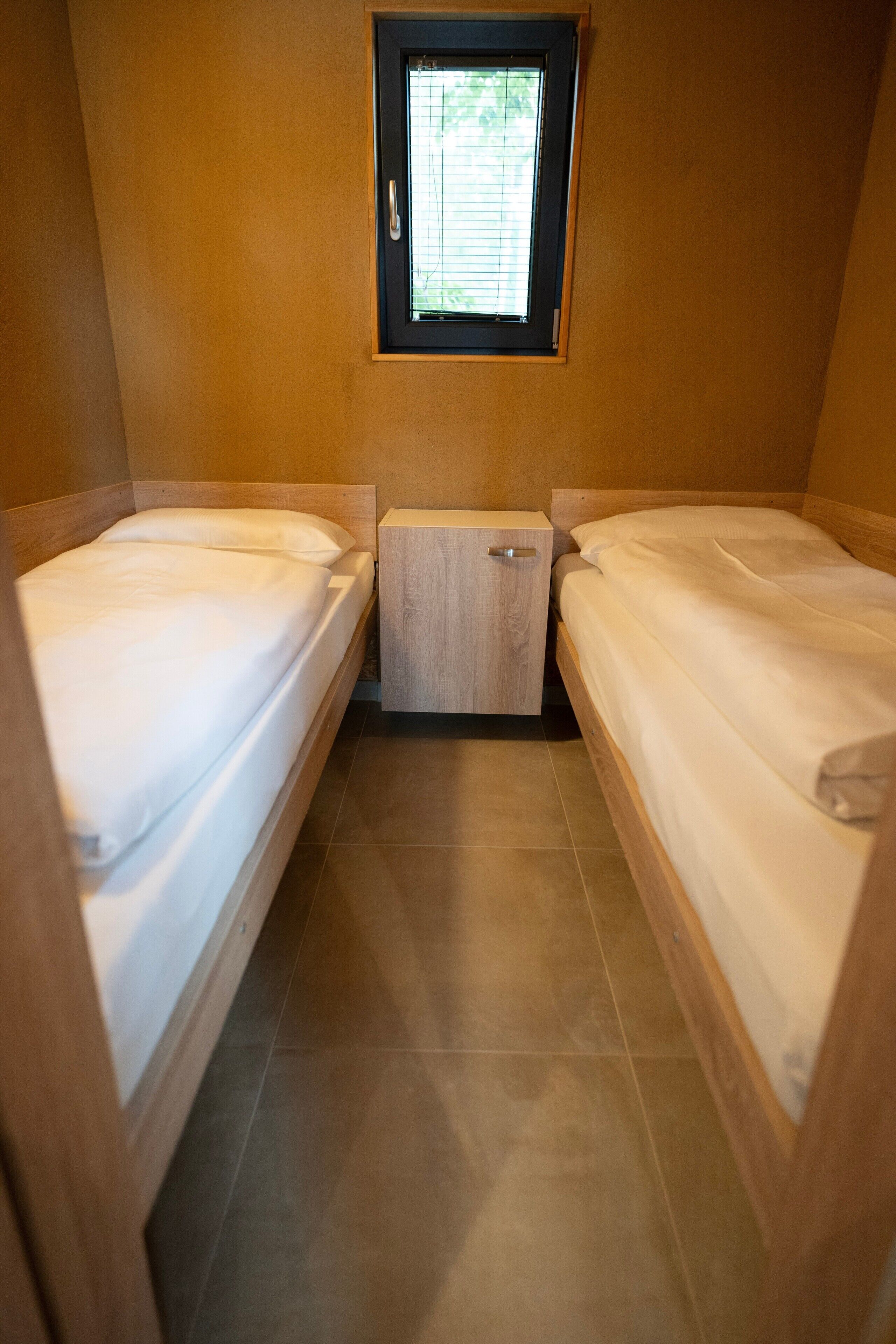 A small, single room with two beds, a window, and a wooden cabinet.
