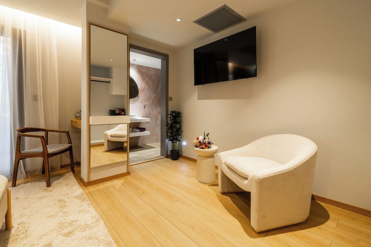 A modern hotel room with a wooden floor, a white armchair, a wooden chair, a small side table with a vase of flowers, a mirror, a flat-screen TV mounted on the wall, and a bathroom visible through an open door.