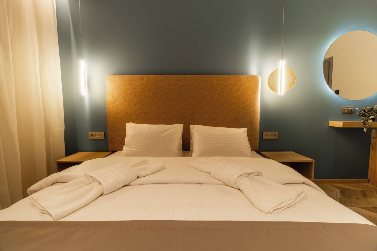 A neatly made bed with white linens and towels, a wooden headboard, bedside tables with lamps, and a round mirror on the wall.