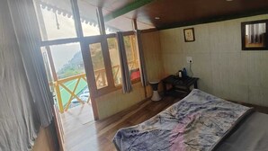 A room with a bed, wooden flooring, a window with curtains, a small table with a chair, and a view of a green landscape outside. - Anany Resort (Pangoot)