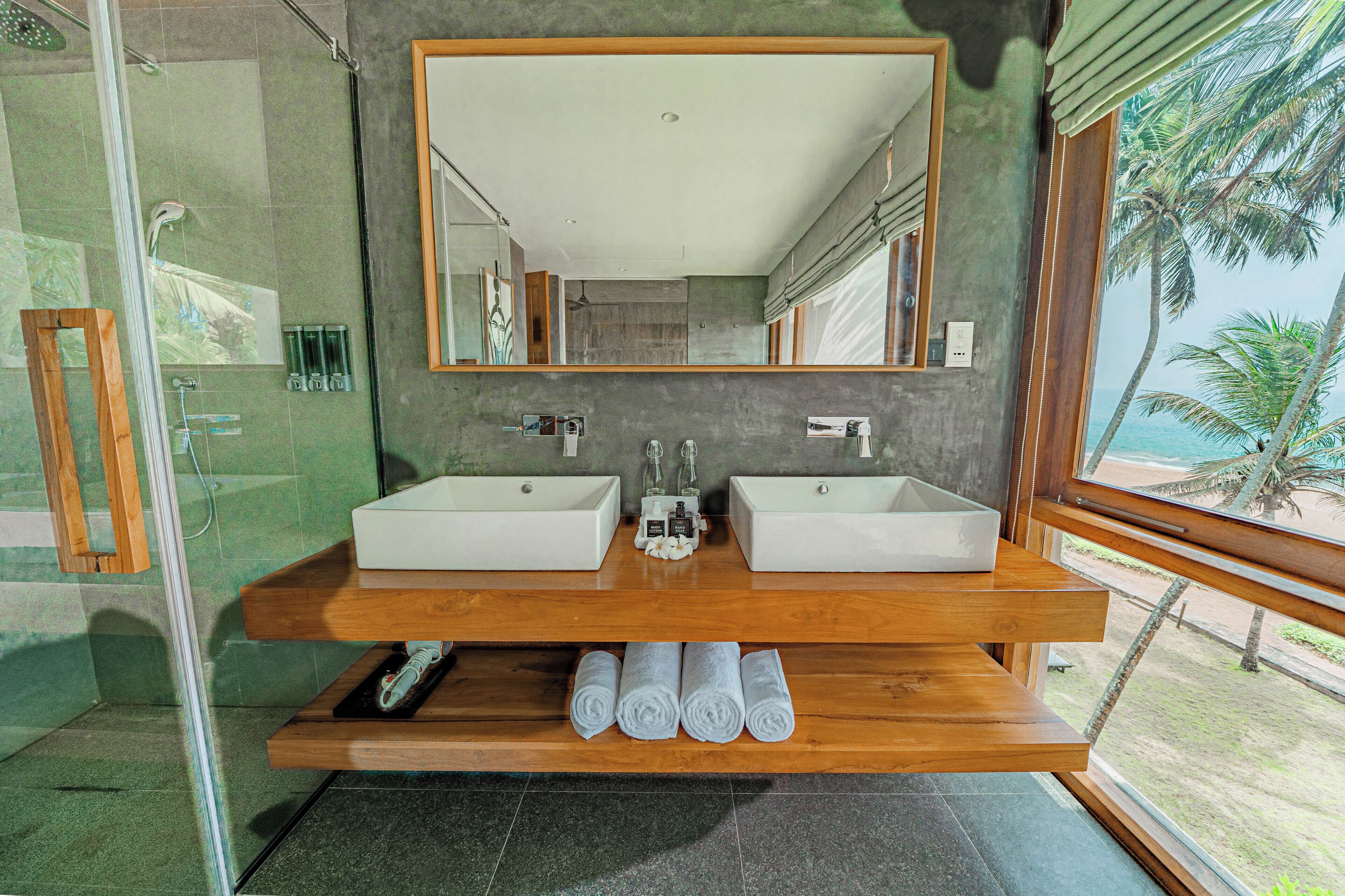 A modern bathroom with two sinks, a large mirror, and a view of the beach.