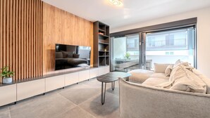 Ein modernes Wohnzimmer mit einem großen Flachbildfernseher an einer Holzwand, einem grauen Sofa, einem kleinen runden Tisch und einer Glastür zum Balkon.