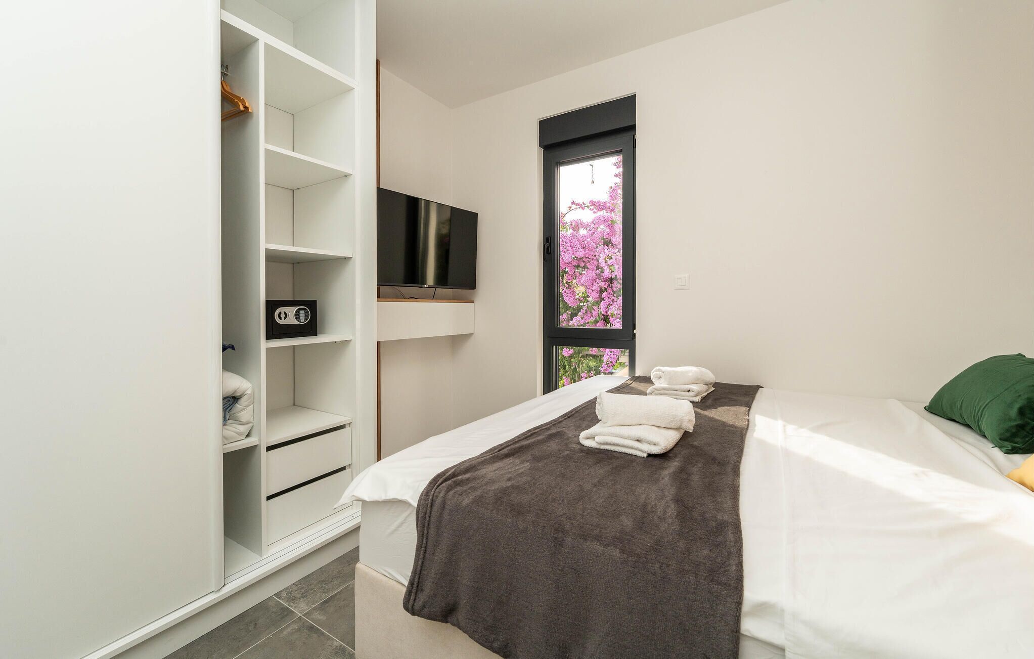Une chambre à coucher avec un lit, un téléviseur fixé au mur, une fenêtre ornée de fleurs roses et une armoire équipée d’une radio.