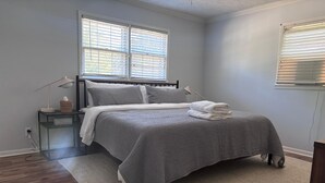 Una cama bien hecha con una colcha gris, una mesita de noche con lámpara y una ventana con persianas.
