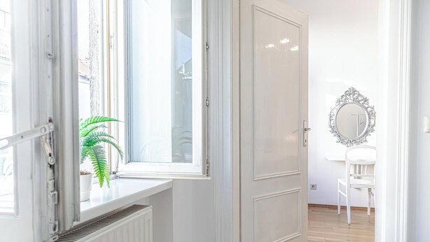 Une pièce avec un radiateur, une fenêtre ornée d’une plante, une porte blanche et une chaise blanche.
