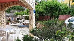 A stone archway leading to a patio with white chairs and a table, surrounded by greenery and a parked car. - Holiday apartment with terrace and air conditioning (Dobrinj)