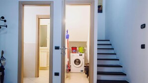 Un pasillo con una lavadora y un cubo rojo en el suelo, que conduce a una escalera.