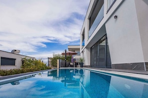 A modern outdoor swimming pool with a clear blue sky and a building with large windows and a flat roof. - Holiday apartment with a beautiful pool (Njivice)