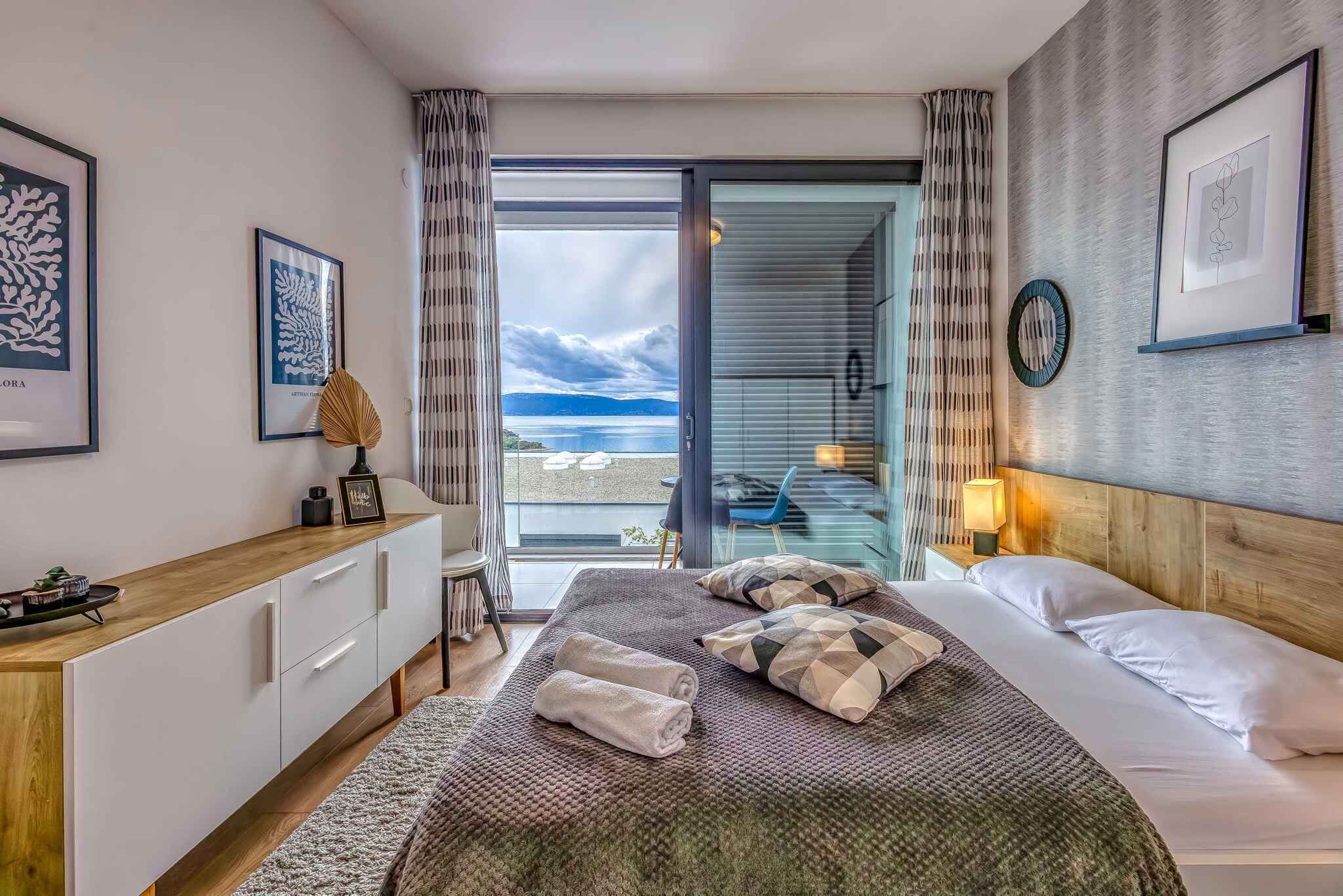 Habitación de hotel moderna con una cama grande, una mesita de noche de madera, un escritorio y vistas al mar a través de una puerta corrediza de vidrio.