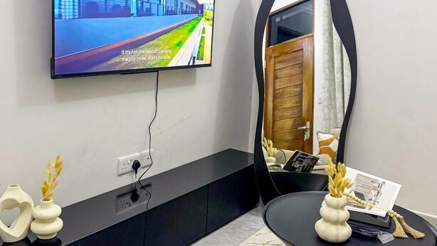 Una sala de estar moderna con un televisor de pantalla plana montado en la pared, una consola negra con objetos decorativos, una mesa de centro redonda negra y un espejo grande que refleja la habitación.