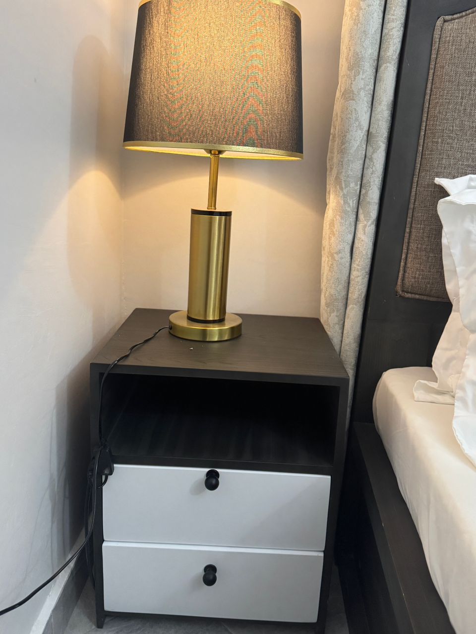 A bedside table with a lamp, a drawer, and a bed with a white pillow.