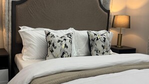 Una cama bien tendida con ropa de cama blanca y almohadas estampadas con motivos florales, un cabecero oscuro y una lámpara de noche.