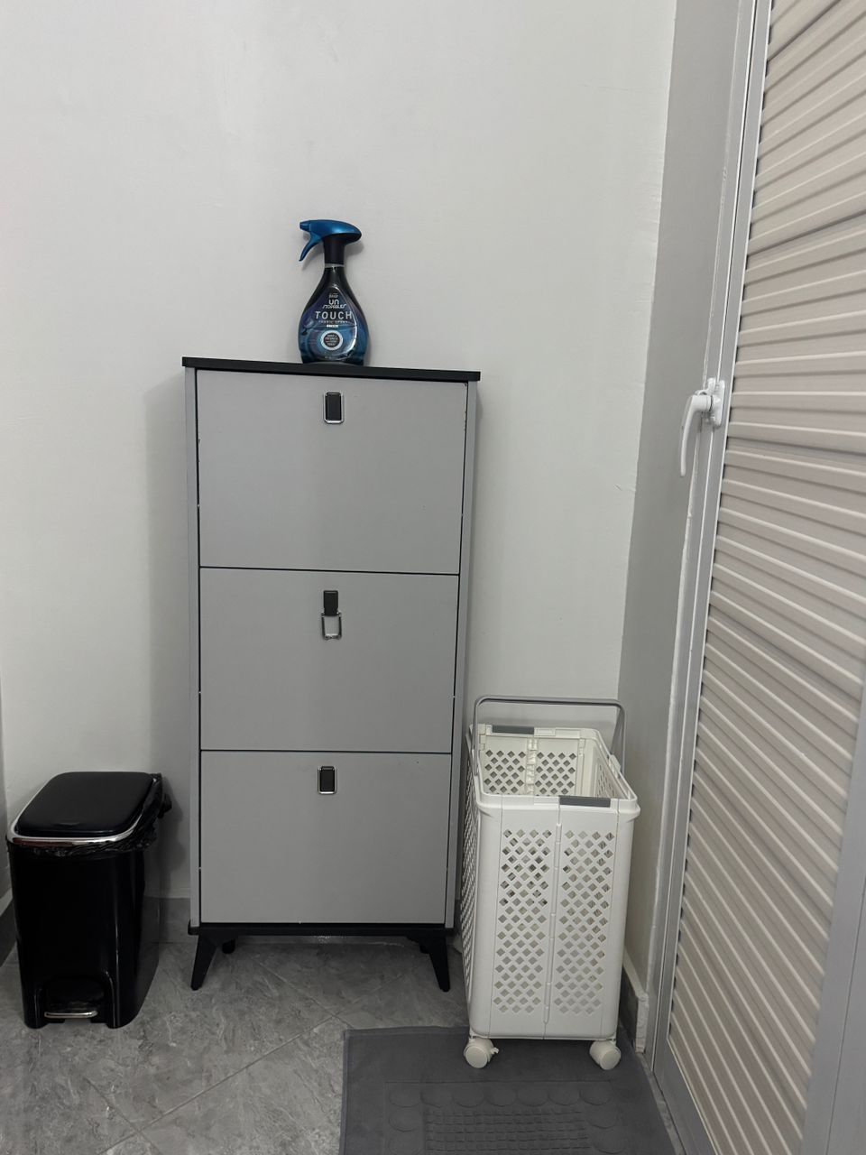 Un pequeño cuarto de servicio con un armario metálico gris, un cesto de basura negro y una cesta de lavandería blanca.