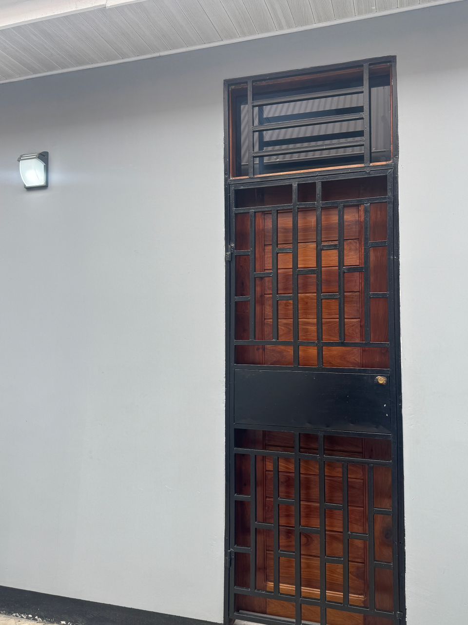 Puerta con una reja metálica y paneles de madera, instalada en una pared lisa con un interruptor de luz arriba.