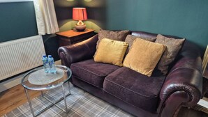 Ein dunkelbraunes Ledersofa mit zwei Kissen, ein kleiner runder Glastisch mit Wasserflaschen, eine Lampe auf einem Holznachttisch und ein karierter Teppich.