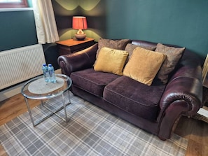 Ein dunkelbraunes Ledersofa mit zwei Kissen, ein kleiner runder Glastisch mit Wasserflaschen, eine Lampe auf einem Holznachttisch und ein karierter Teppich.