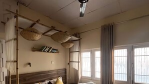 A bedroom with a bed, ceiling fan, windows, and a shelf with books. - Summer valley cottage (Mant Khas)