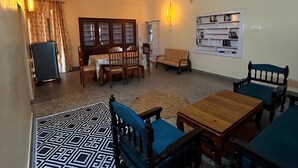 A spacious room with a high ceiling, a ceiling fan, a wall-mounted shelf with books, a patterned rug, and a dining table with chairs. - Summer valley cottage (Mant Khas)