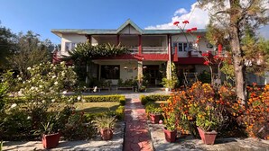 A two-story building with a central porch, surrounded by well-maintained gardens and potted plants. - Summer valley cottage (Mant Khas)