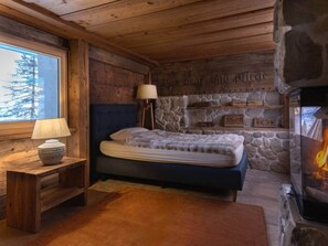 A cozy log cabin bedroom with a stone fireplace, a bed with a stone headboard, and a window overlooking a snowy landscape.