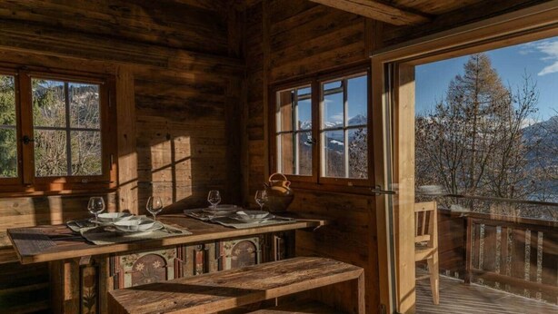 A rustic wooden cabin with a dining area, large windows, and a view of a lake and mountains.