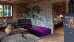 A rustic cabin interior with a stone wall, wooden ceiling, a sofa, a purple ottoman, a fireplace, and a window with a view of trees.