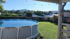 An outdoor swimming pool area with a covered patio, string lights, and a view of houses and mountains in the background. - The Wonky House (Bowraville)