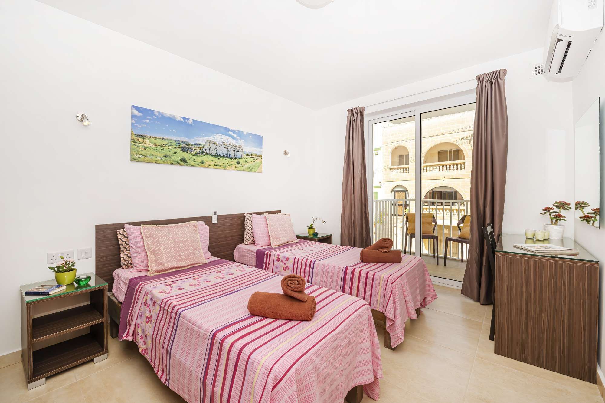 Una habitación de hotel con dos camas, un balcón con vista a un edificio, un cuadro en la pared y una mesita con una planta.