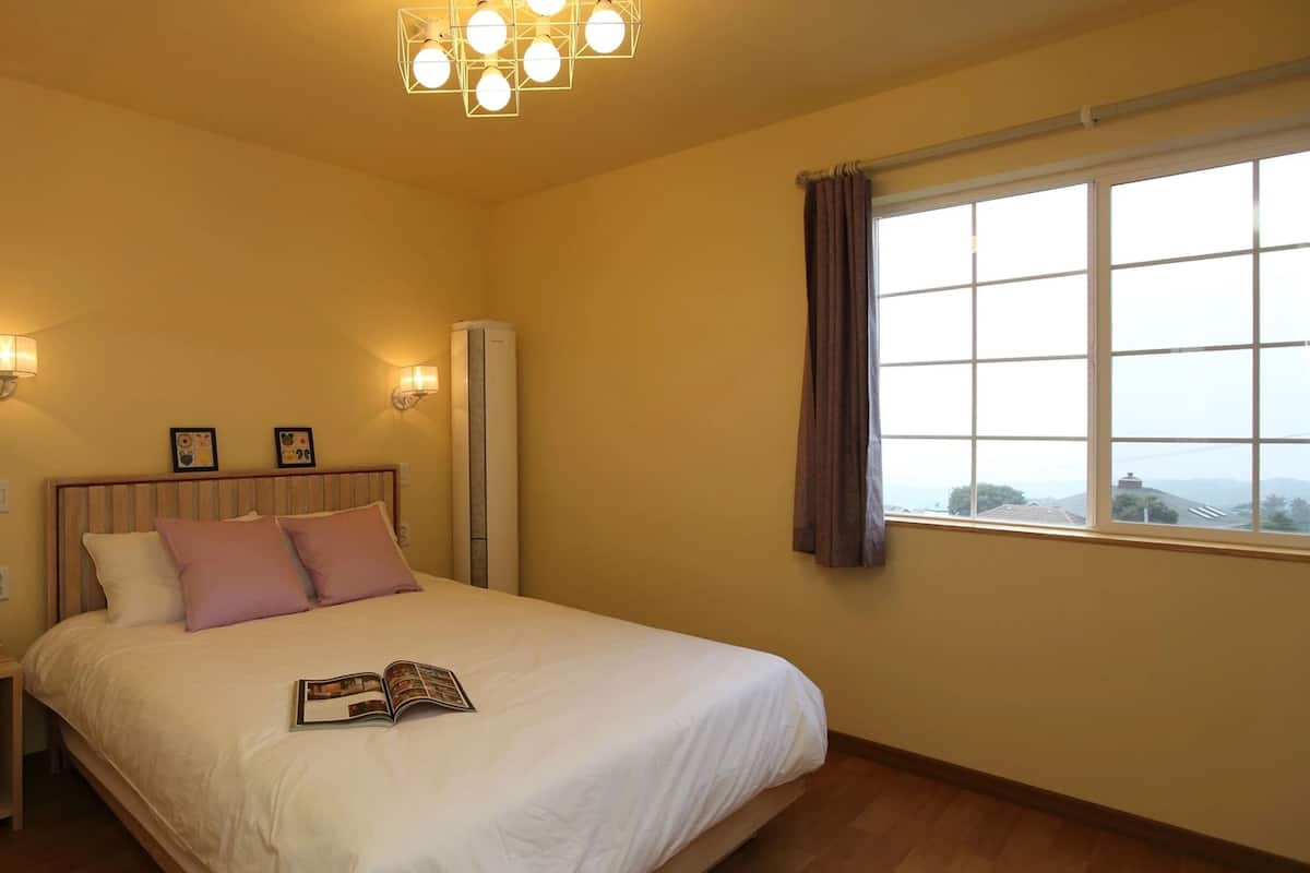 Un dormitorio con cama, lámparas de noche, una mesita de noche con una planta, ventana con cortinas y aire acondicionado de pared.