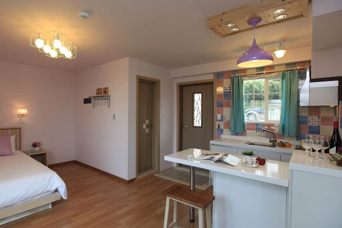 Un dormitorio con cama, mesita de noche y una pequeña mesa con una silla. Hay una puerta que da a un baño y una kitchenette con ventana.