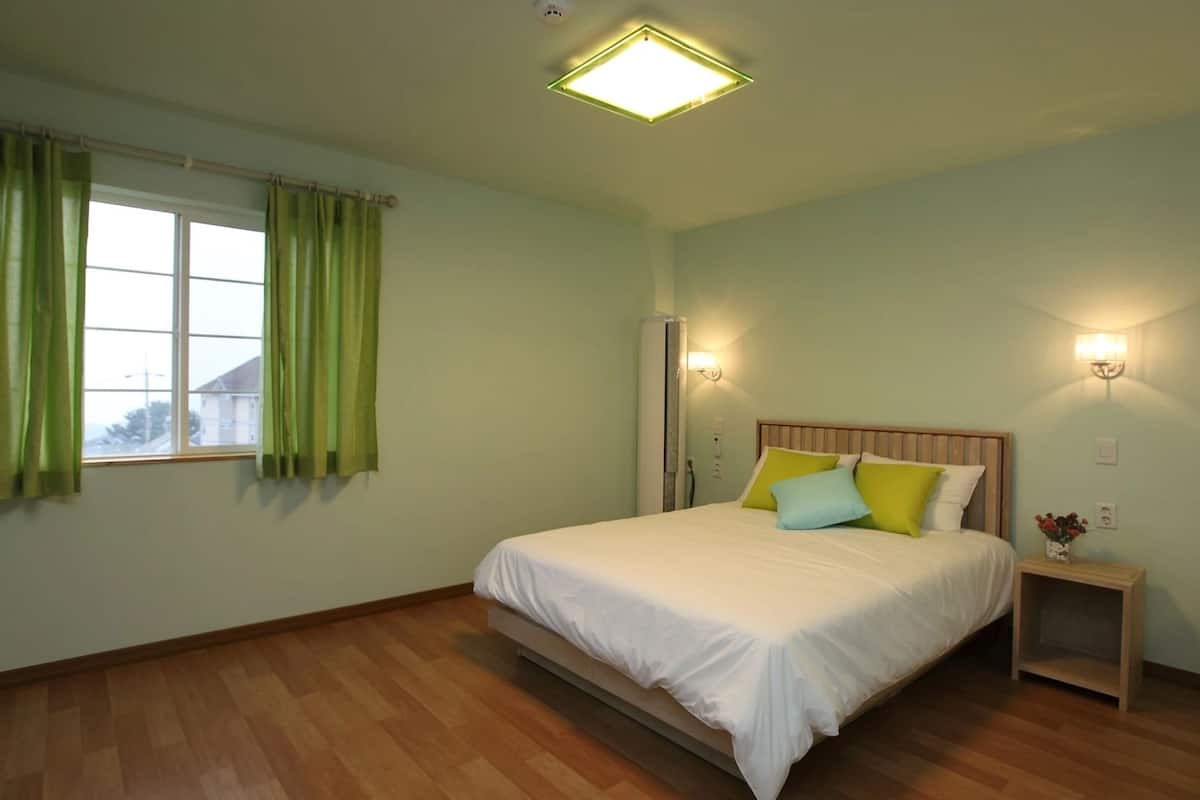 Un dormitorio con cama, cortinas verdes, una mesita de noche con lámpara y una luz de techo.