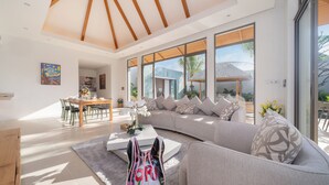 A modern living room with a curved sofa, a dining area, and large windows offering a view of the outdoors. - Tropical Pool Anchan Indigo 16 (Thep Krasattri)
