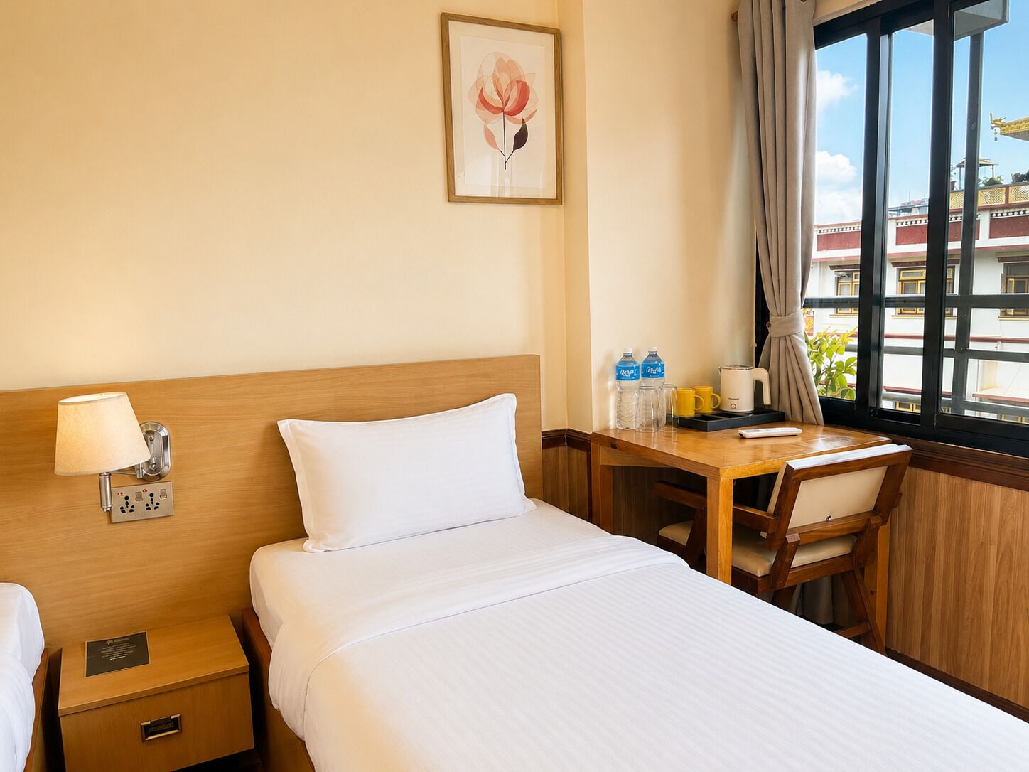 A hotel room with a bed, bedside table, lamp, and a window with a view of buildings.