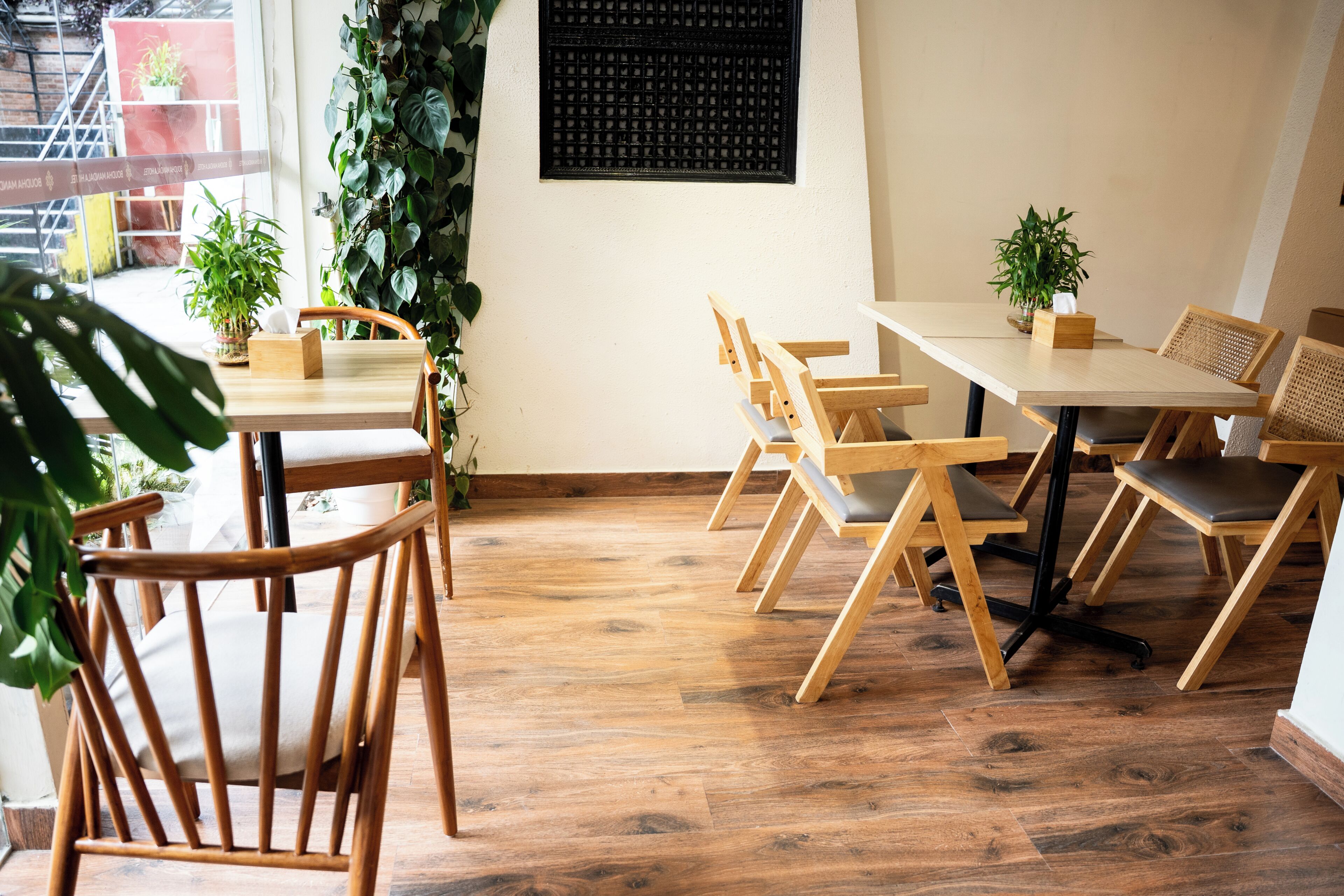 Un coin repas en bois avec des tables et des chaises, un mur noir orné d’un motif quadrillé et des plantes vertes.
