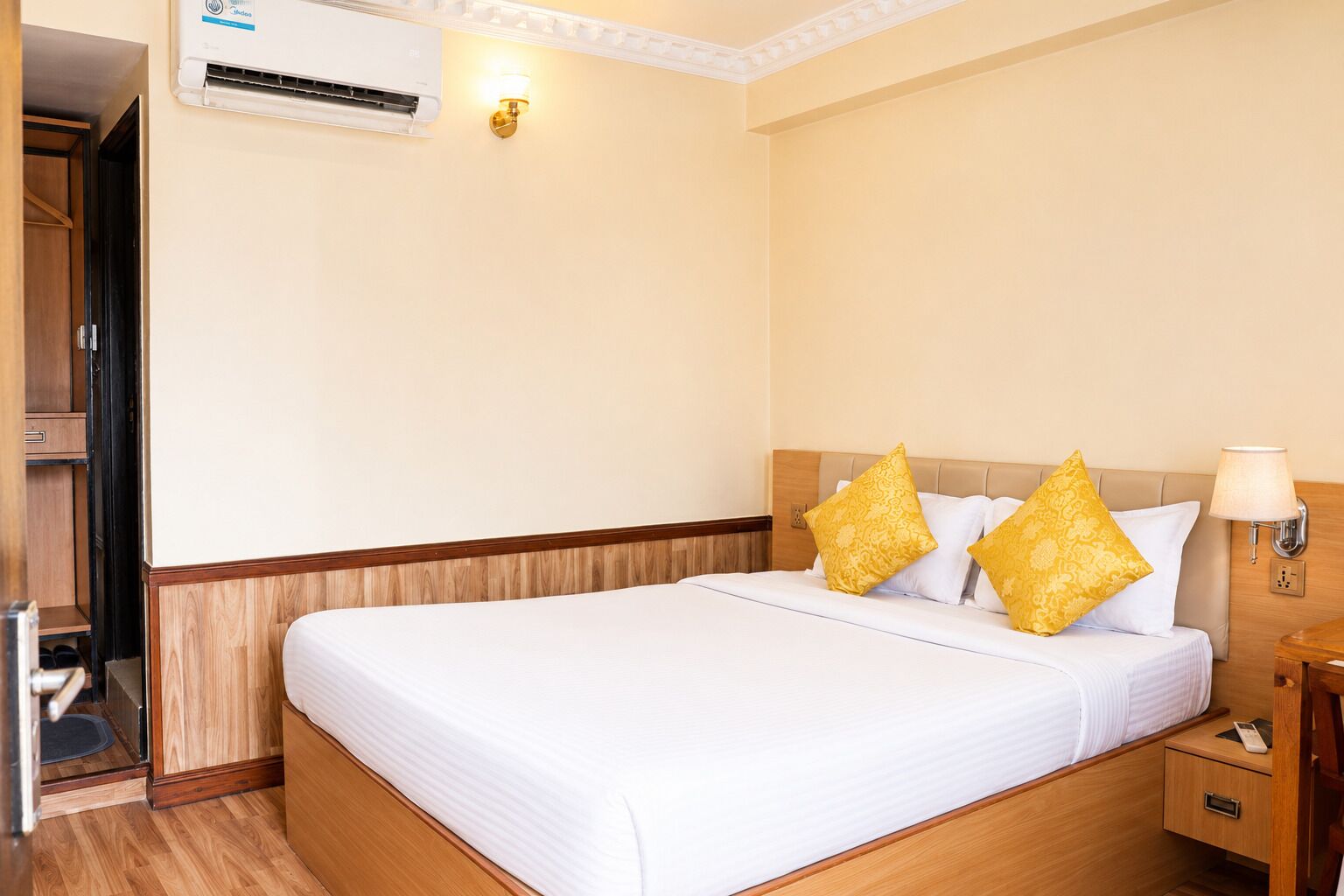 A hotel room with a bed, a nightstand, a lamp, an air conditioning unit, and a wall-mounted light fixture.