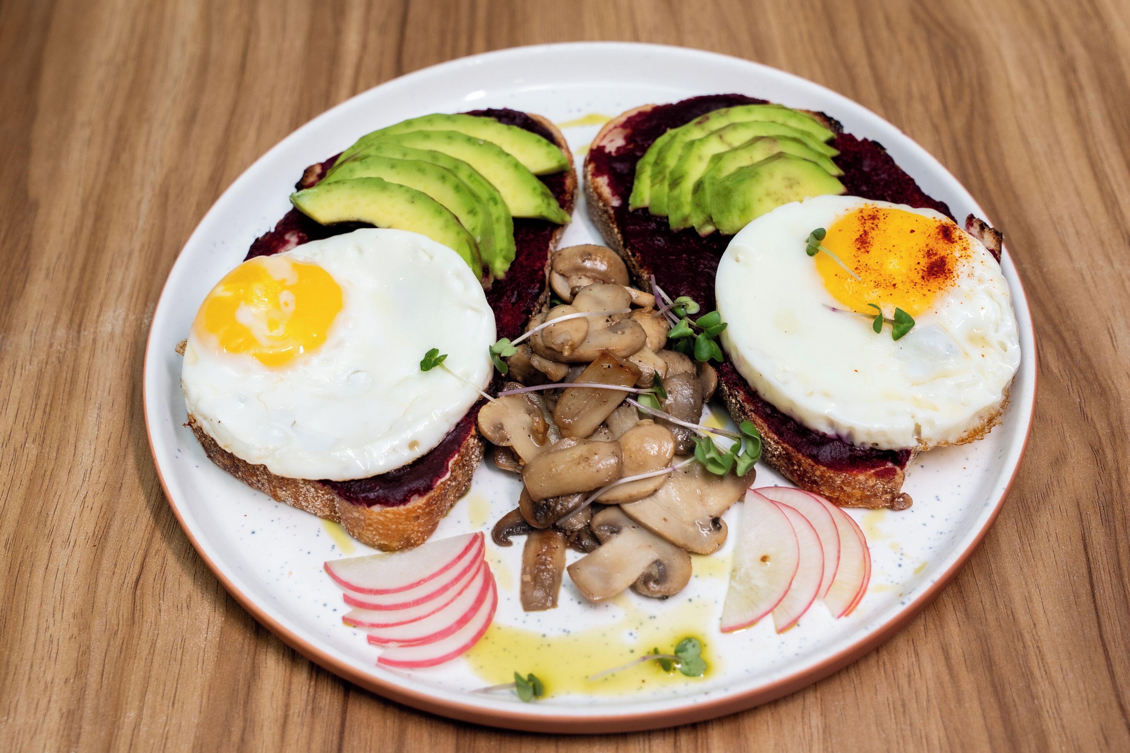 Une assiette avec deux sandwichs ouverts, chacun garni d’un œuf au plat, de tranches d’avocat et accompagné d’une portion de champignons.