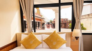 Quarto de hotel com uma cama grande, duas almofadas decorativas, uma janela com cortinas e vista para a rua.
