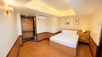 A hotel room with a bed, bedside table, lamp, wardrobe, and an air conditioning unit.