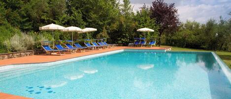 A rectangular swimming pool with clear blue water, surrounded by a tiled deck. There are blue lounge chairs and white umbrellas along the poolside.