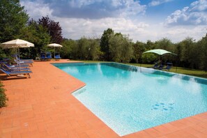 Ein rechteckiger Swimmingpool mit klarem, blauem Wasser, umgeben von einer Ziegelterrasse. Mehrere blaue Liegestühle und Sonnenschirme bieten Schatten.