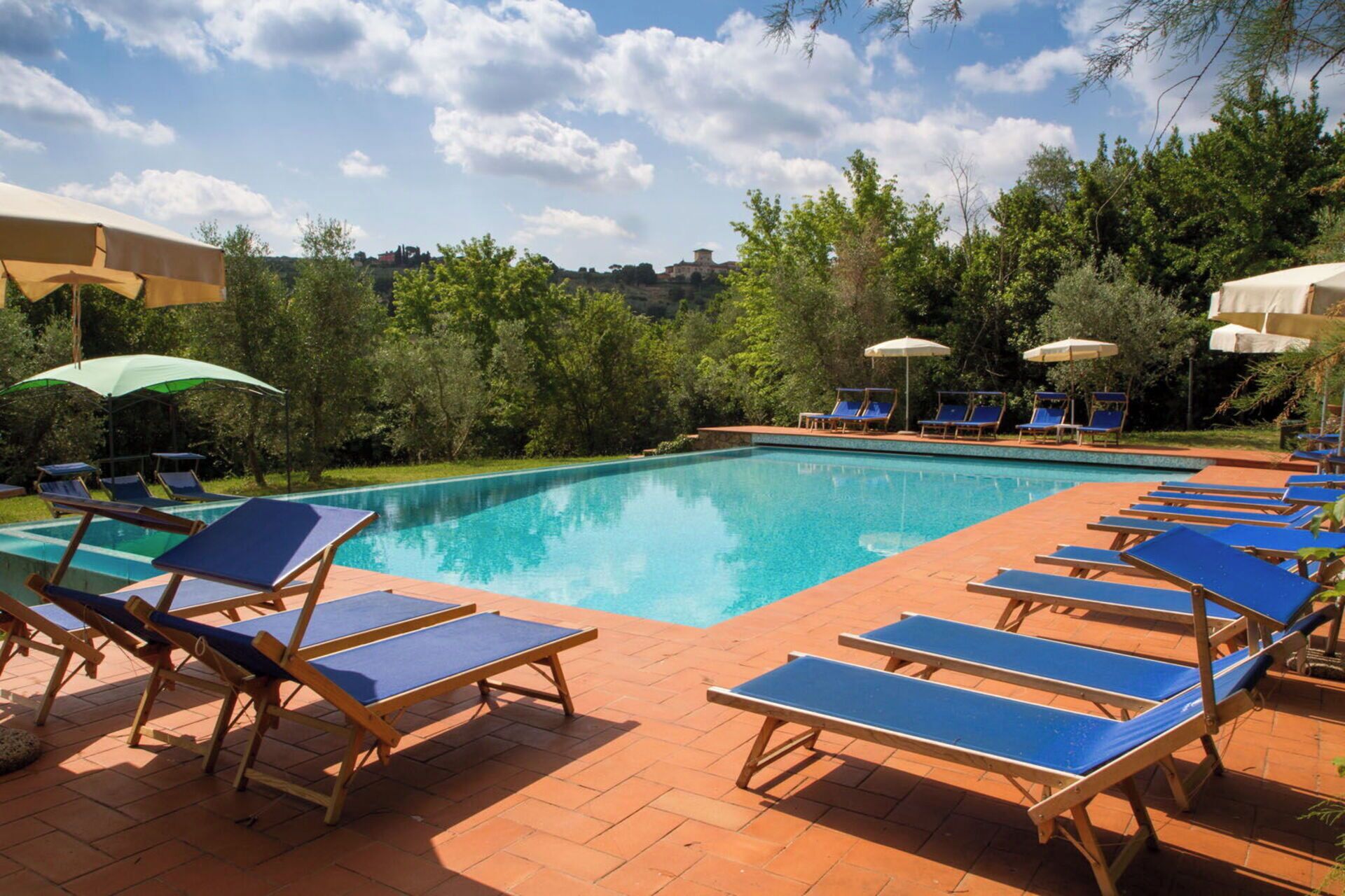A swimming pool with blue lounge chairs and umbrellas, surrounded by trees and a clear sky.