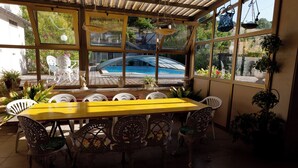Eine überdachte Terrasse mit einem gelben Tisch und verzierten Stühlen, große Fenster mit Blick auf ein Schwimmbad und Grünflächen.