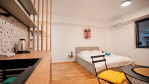 Quarto compacto com cama, uma mesinha, uma cadeira, chaleira e janela.