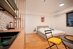 Ein kleines Zimmer mit einem Bett, einem Tisch, einem Stuhl, einem Wasserkocher und einem Fenster.