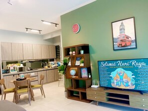Ein modernes Hotelzimmer mit Küchenbereich, einem Esstisch mit Stühlen, einem Bücherregal, einer Uhr und einem an der Wand befestigten Fernseher, der eine Speisekarte zeigt.