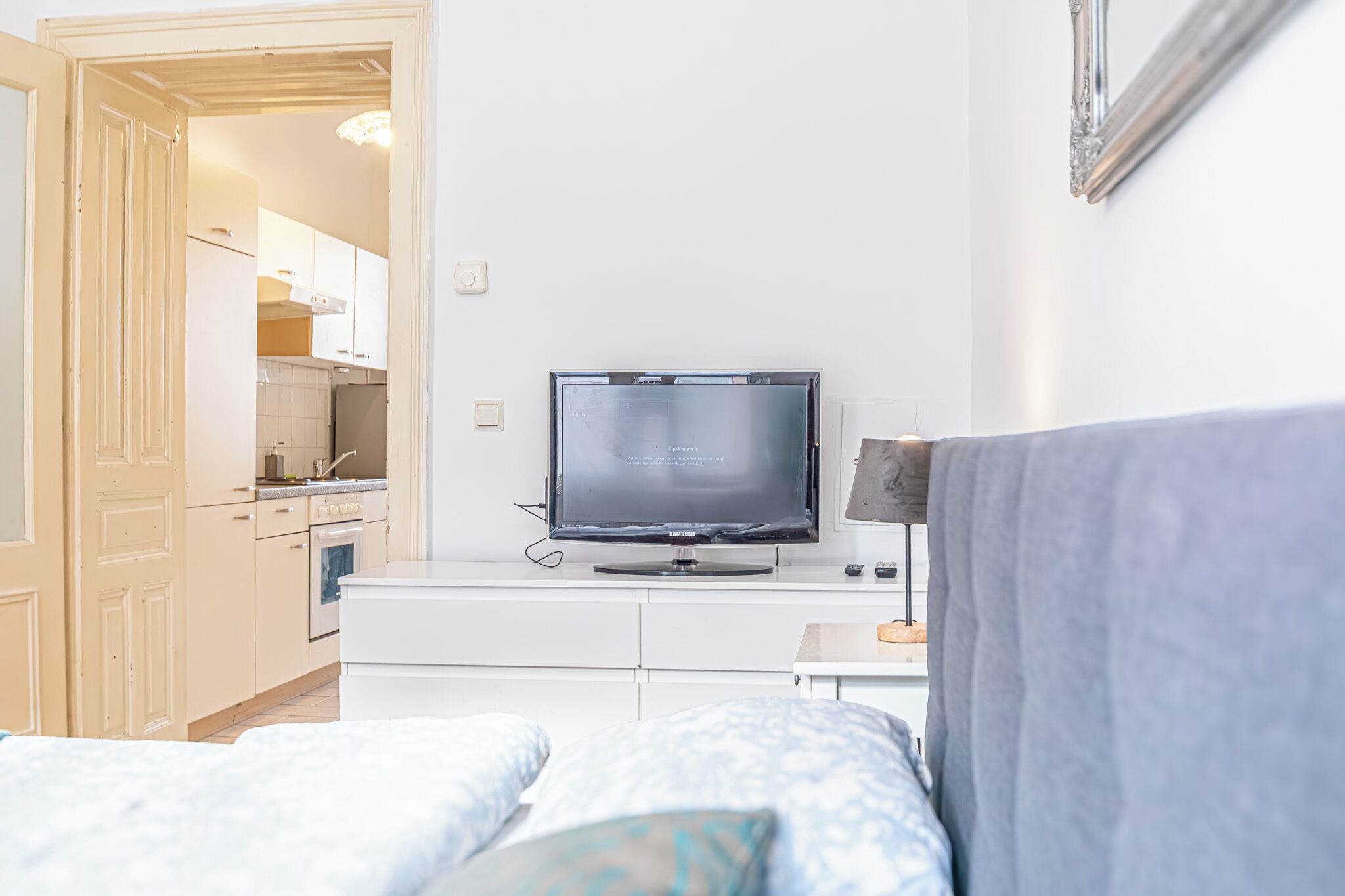 A hotel room with a bed, a TV on a stand, a small table, and a kitchenette in the background.