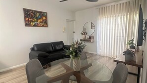 Een moderne woonkamer met een eettafel met glazen blad, grijze stoelen, een zwarte leren bank, een plafondventilator, een schilderij en een raam met dunne gordijnen.