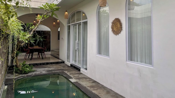 A pool area with a clear view of the pool, a dining area with chairs and a table, and a covered walkway with hanging lights and a decorative wall piece. - Ingrid Villa Ubud by AGATA (Ubud)